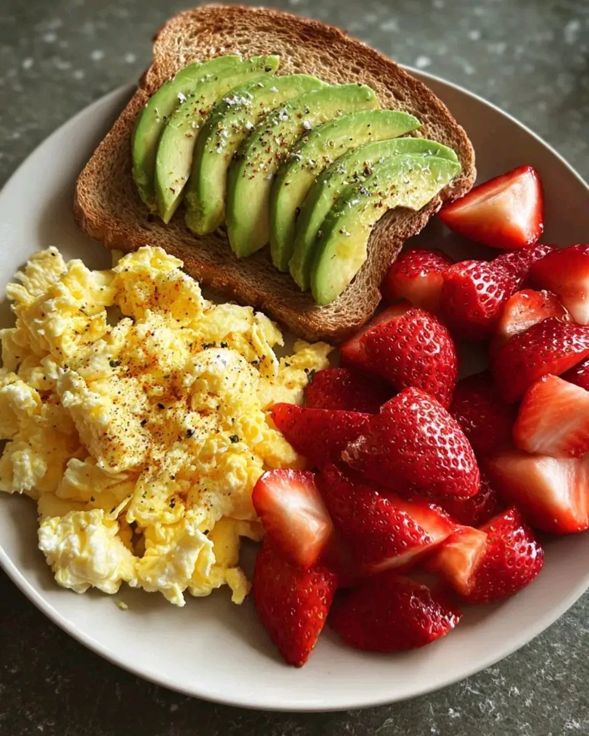 Avocado toast with scrambled eggs and sliced strawberries on a white plate.