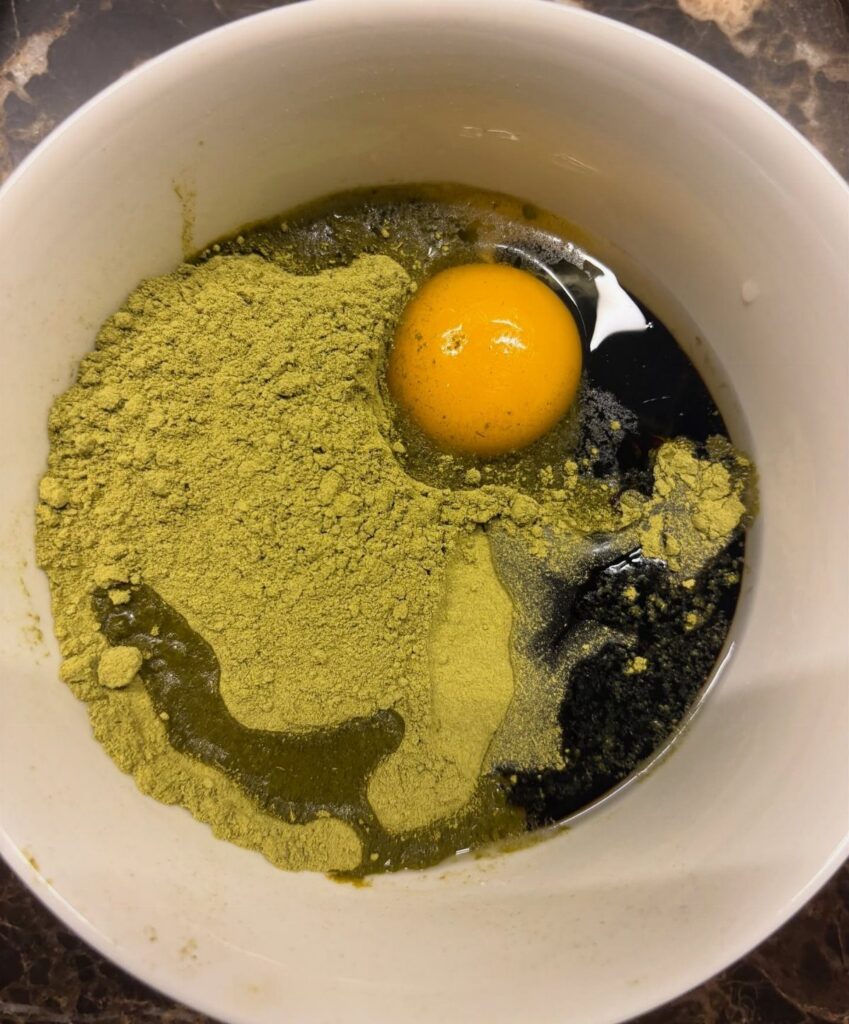 Raw egg yolk and green powder with oil in a white bowl.