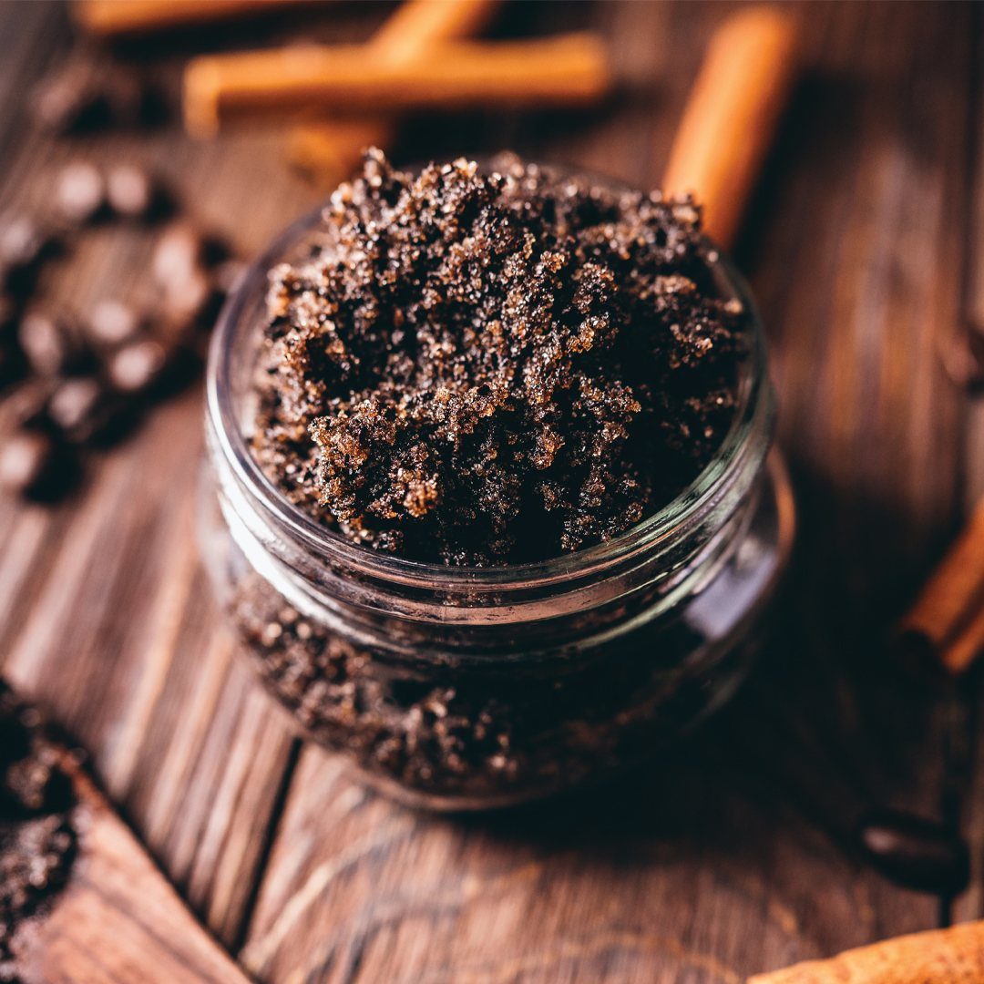 Dark brown granular scrub in a glass jar surrounded by cinnamon and coffee beans.
